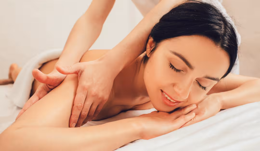 A woman lying on her bed with eyes closed, relaxed and nearly asleep, as a therapist massages her shoulders, showing how a massage for sleep eases you down at home