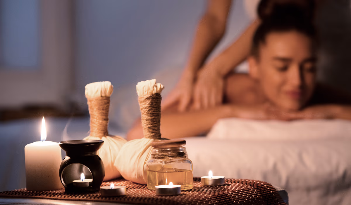 A woman relaxing during a candlelit massage with herbal compress balls and an oil warmer in the foreground, comparing the calm ritual of a home massage vs a spa visit