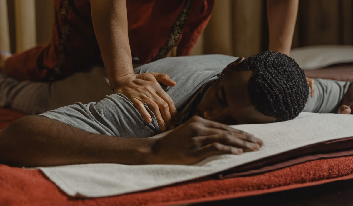 A man lying face down on soft towels in a warmly lit, calm room, eyes closed and relaxed, as a therapist presses his shoulder, showing a home prepared as a calm space for a massage