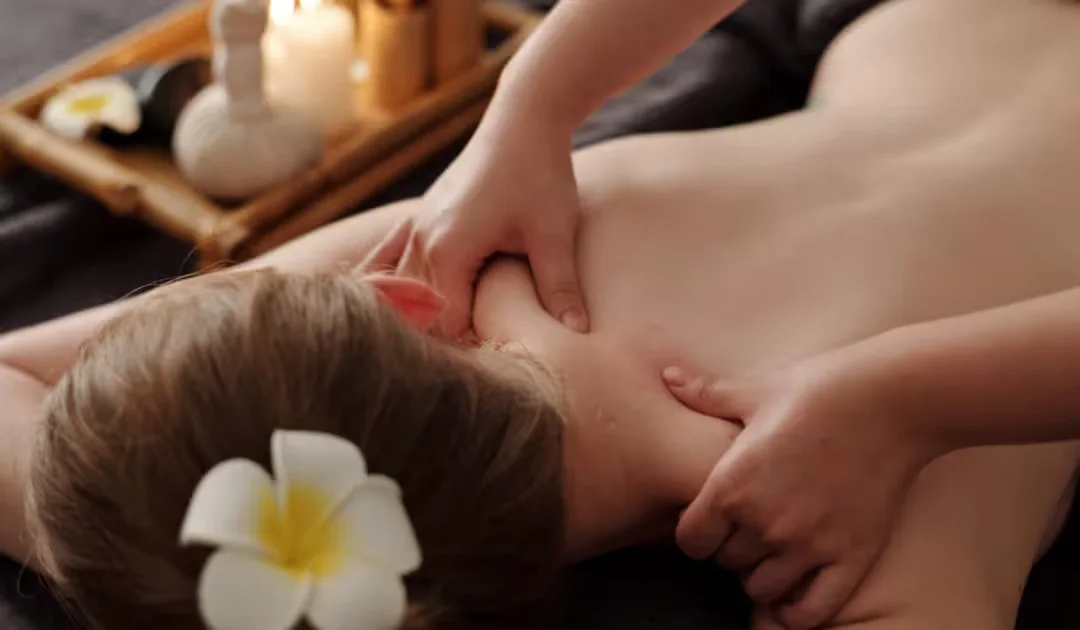 A therapist's hands working a client's neck and shoulders during a relaxing home massage, with a white frangipani flower and soft candlelight nearby